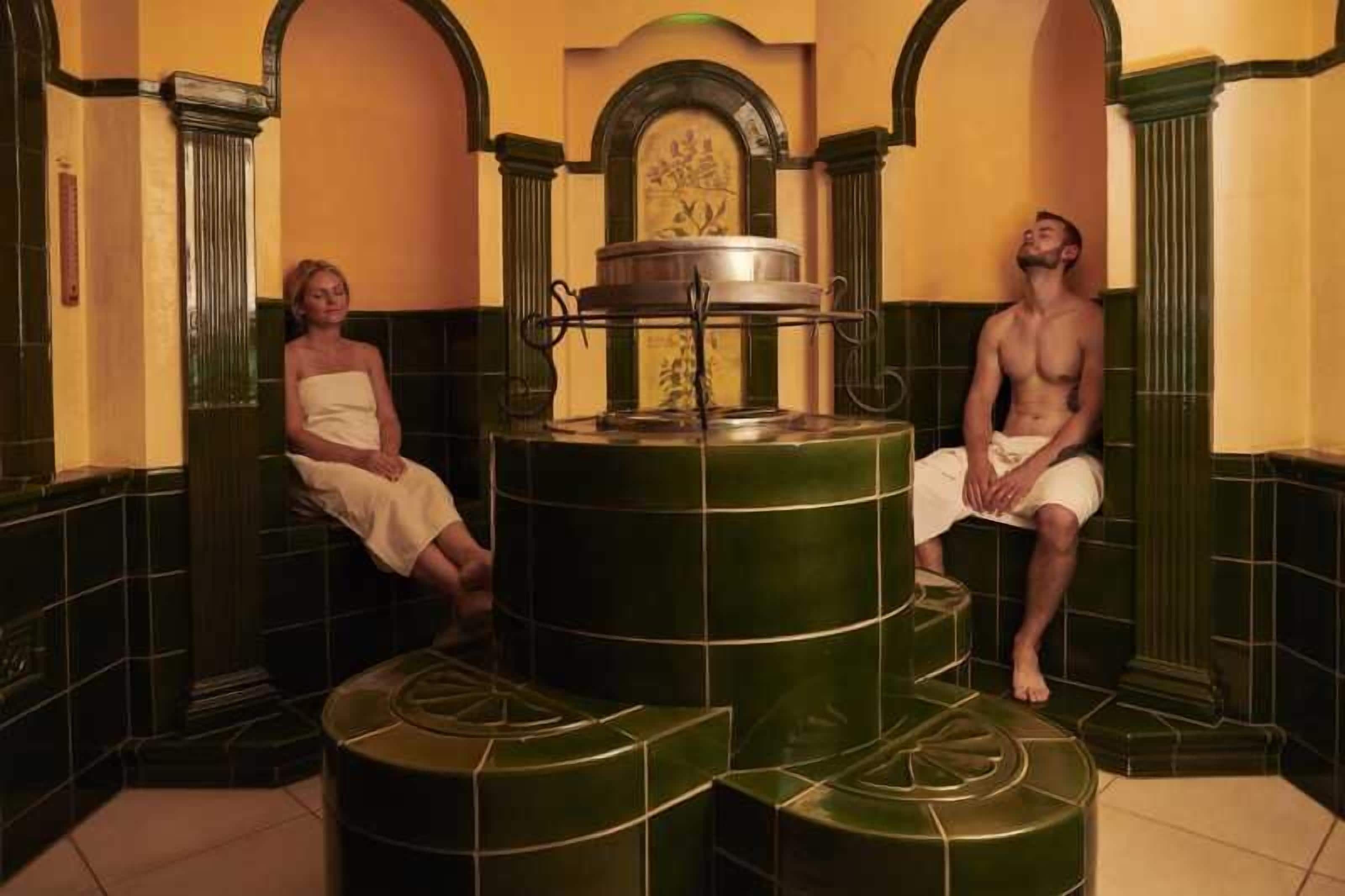 Ein Spa-Bereich mit einem zentralen Dampfbad, zwei entspannende Personen und einer Wand mit floralem Dekor.