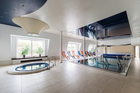Una piscina interior con barrera de vidrio, sillas de descanso y una lámpara en el techo.