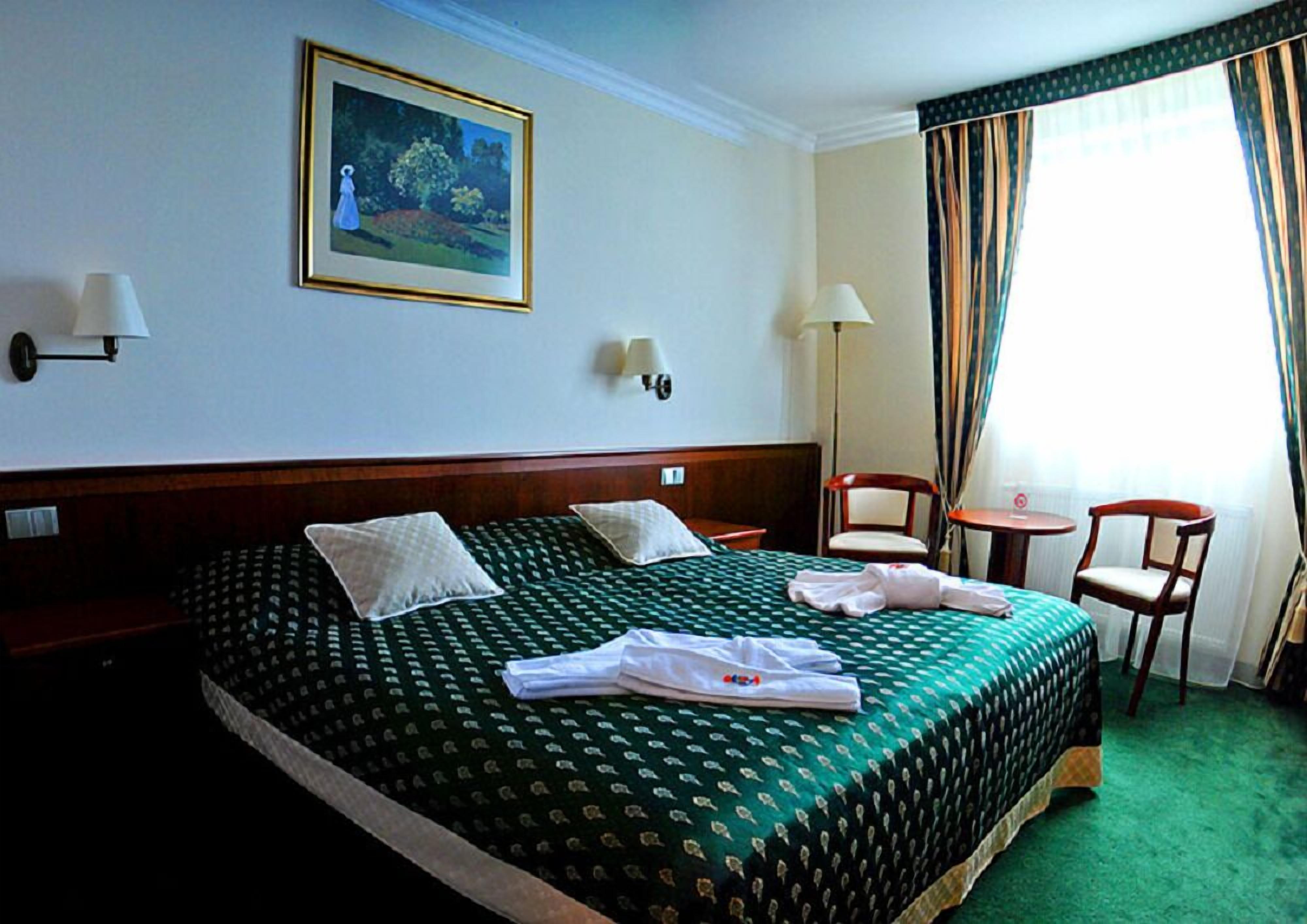 A hotel room with a large bed, two bedside lamps, a painting on the wall, a small table with chairs, and a window with curtains.