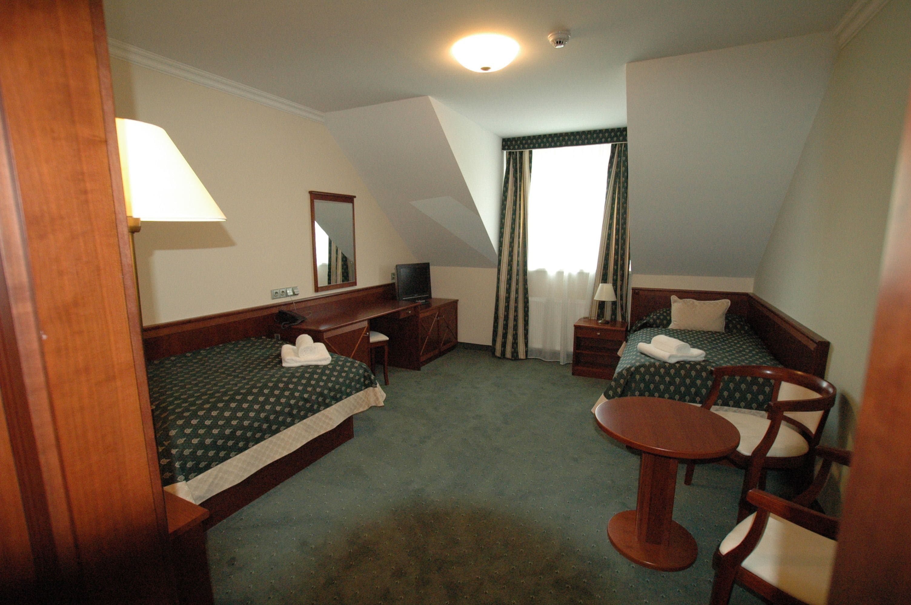 A hotel room with two single beds, a desk, a chair, a small table, a television, and a window with curtains.