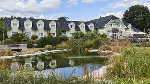 A hotel with a pond in front, surrounded by greenery and a clear sky. - Hotel Komorni Hurka (Cheb)