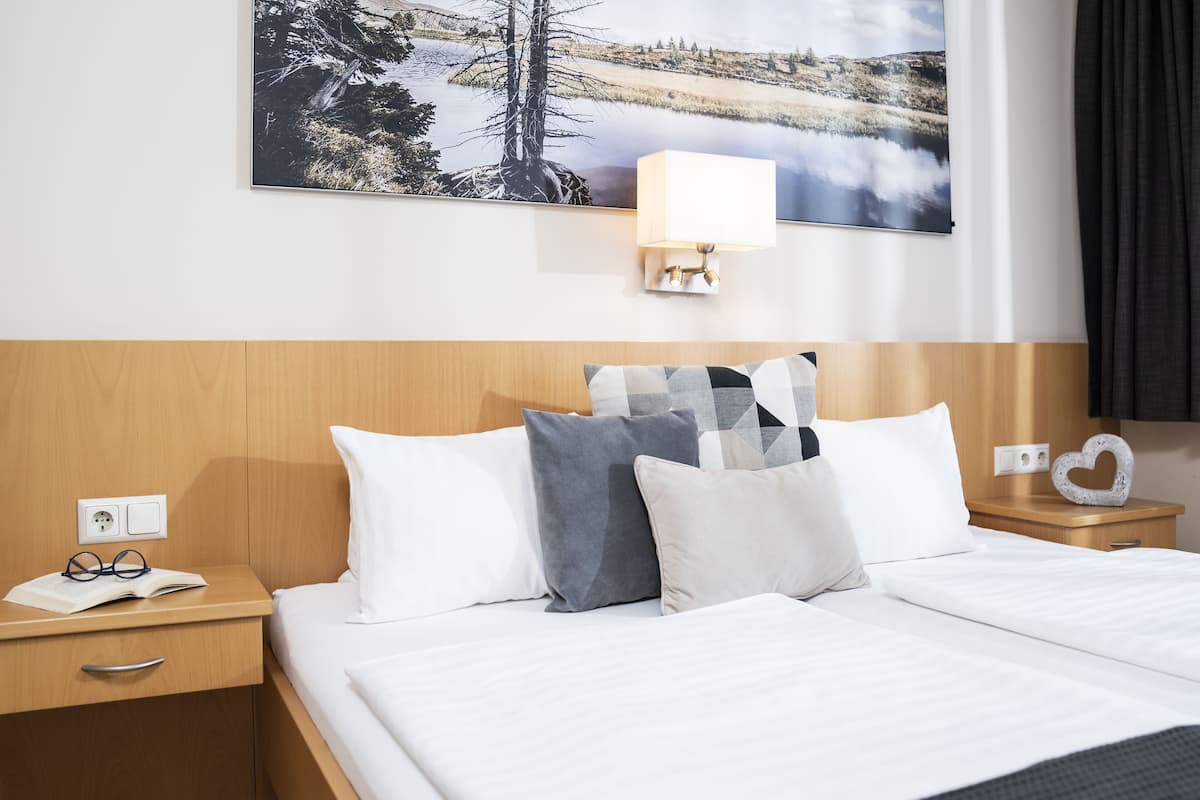 Quarto de hotel com uma cama, duas mesas de cabeceira, uma luminária na parede e um quadro grande com uma paisagem.