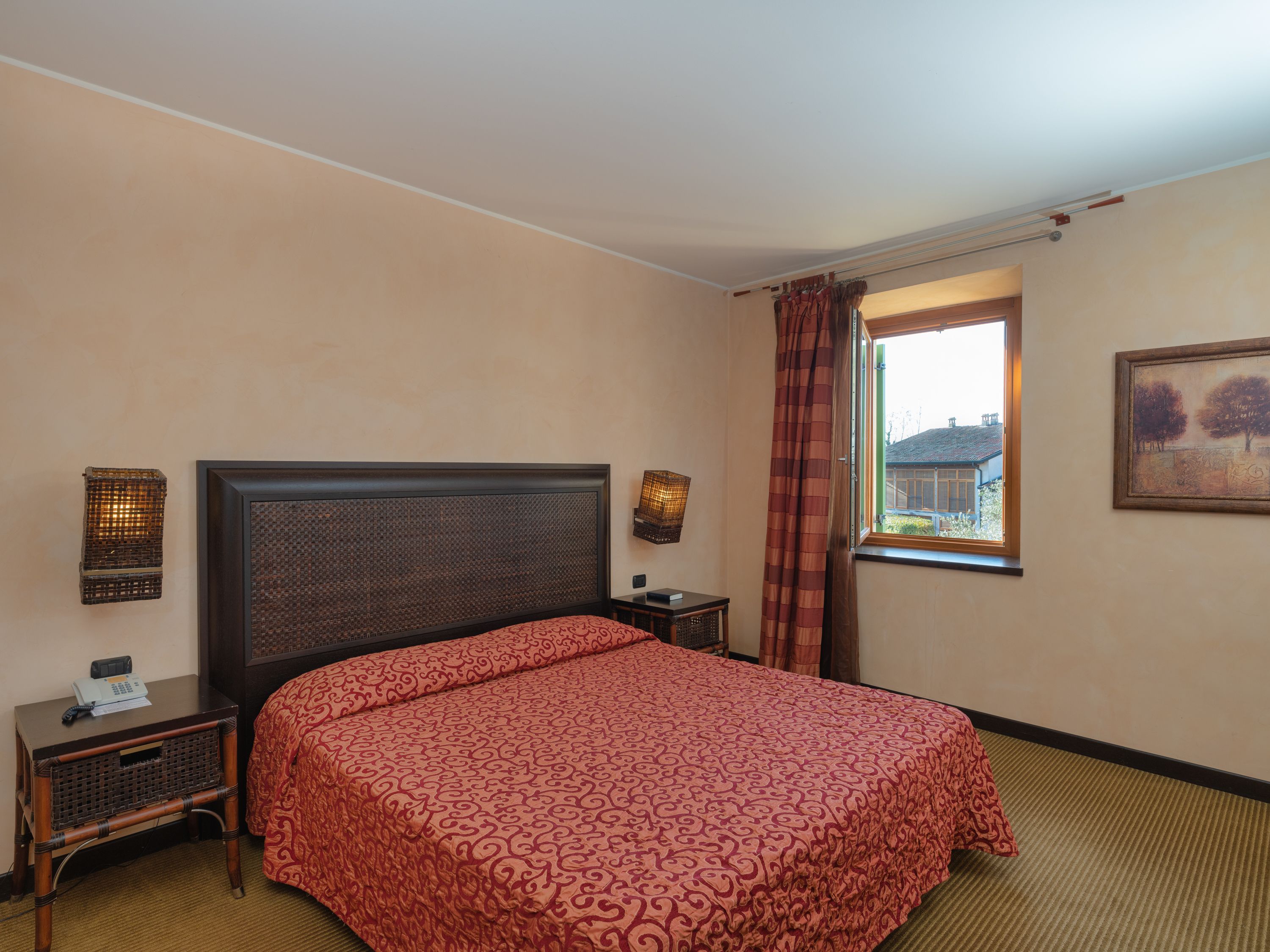 A hotel room with a large bed, a bedside table with a lamp, a window with curtains, and a painting on the wall.