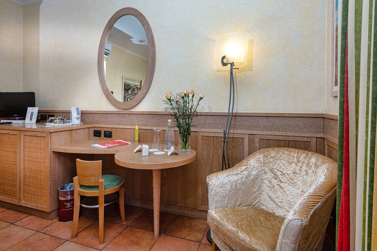 A hotel room with a wooden desk, a chair, a round table, a mirror, a floor lamp, and a large cushioned armchair.