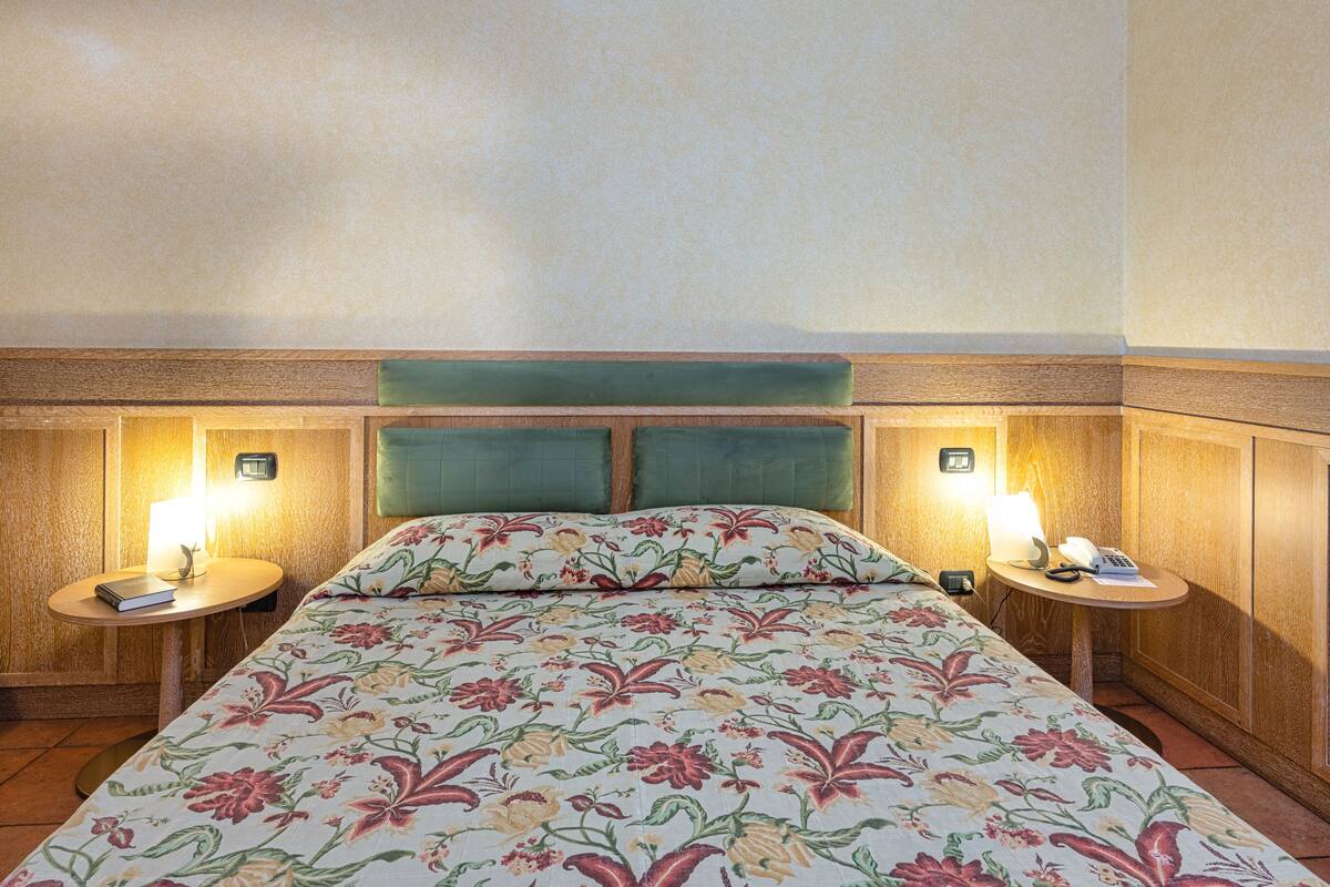 A hotel room with a floral bedspread, wooden headboard, two bedside tables with lamps, and a small round table with a telephone and a book.