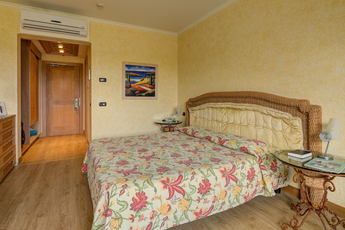 A bedroom with a floral bedspread, a wooden door, a painting on the wall, and a bedside table with a lamp.