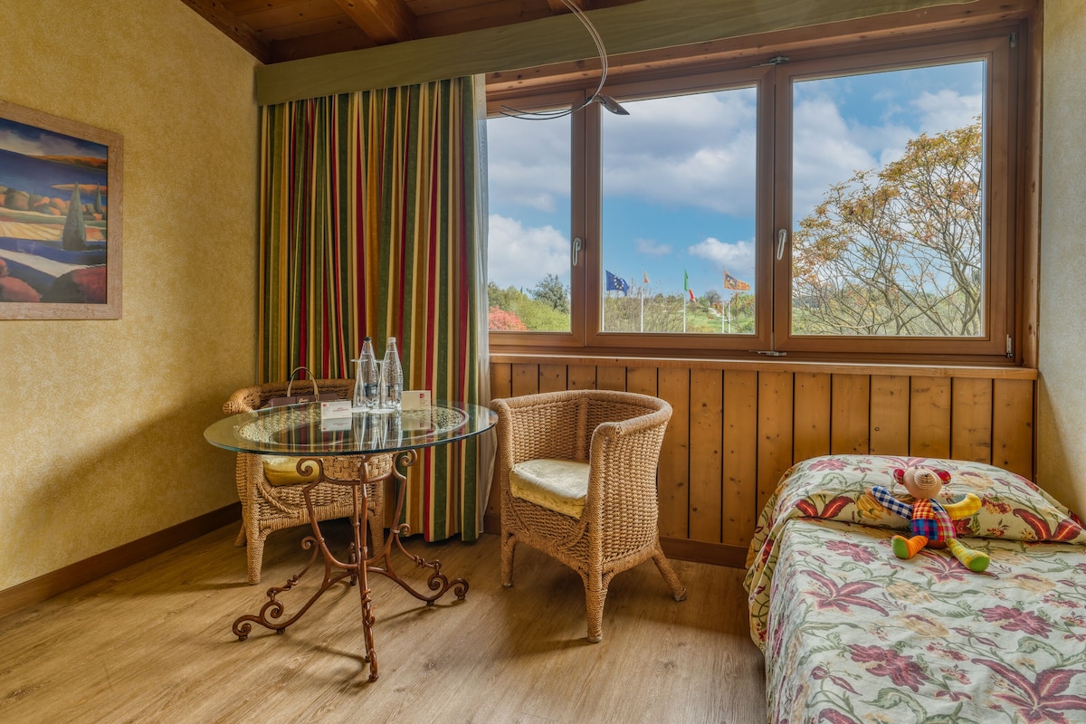 A cozy room with a wooden ceiling, a large window with a view of trees and flags, a wicker chair, a glass table with bottles, and a bed with a floral bedspread.