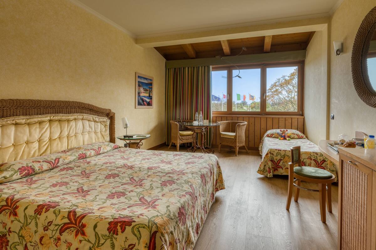 A room with two beds, a dining table, chairs, a small table with a lamp, and a window with flags.