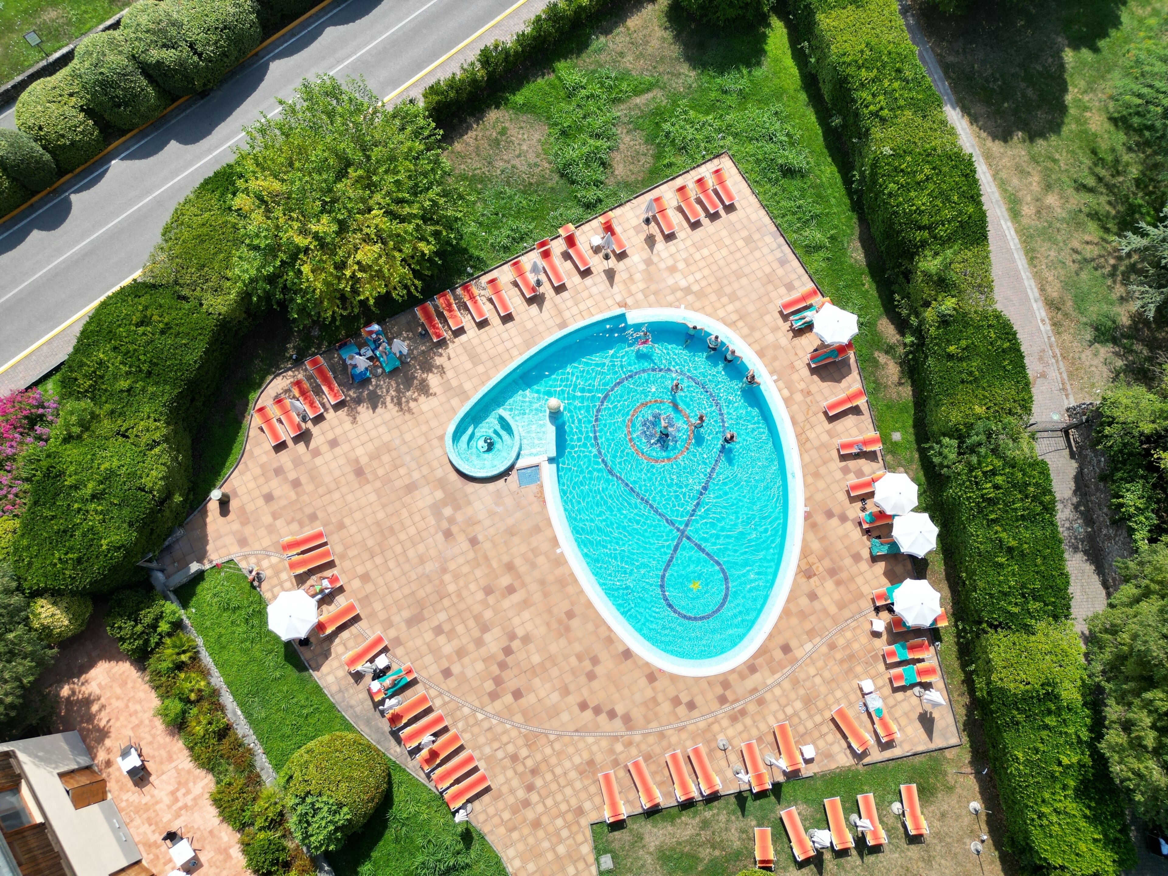 Une vue aérienne d’une piscine entourée de chaises longues et de parasols, avec un chemin pavé et de la végétation.