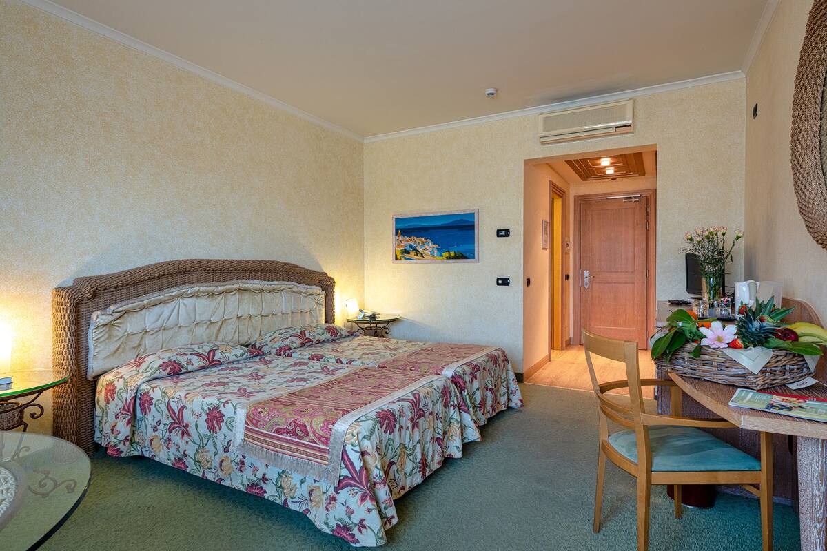 A hotel room with a bed, a chair, a table with a basket of fruit, and a painting on the wall.