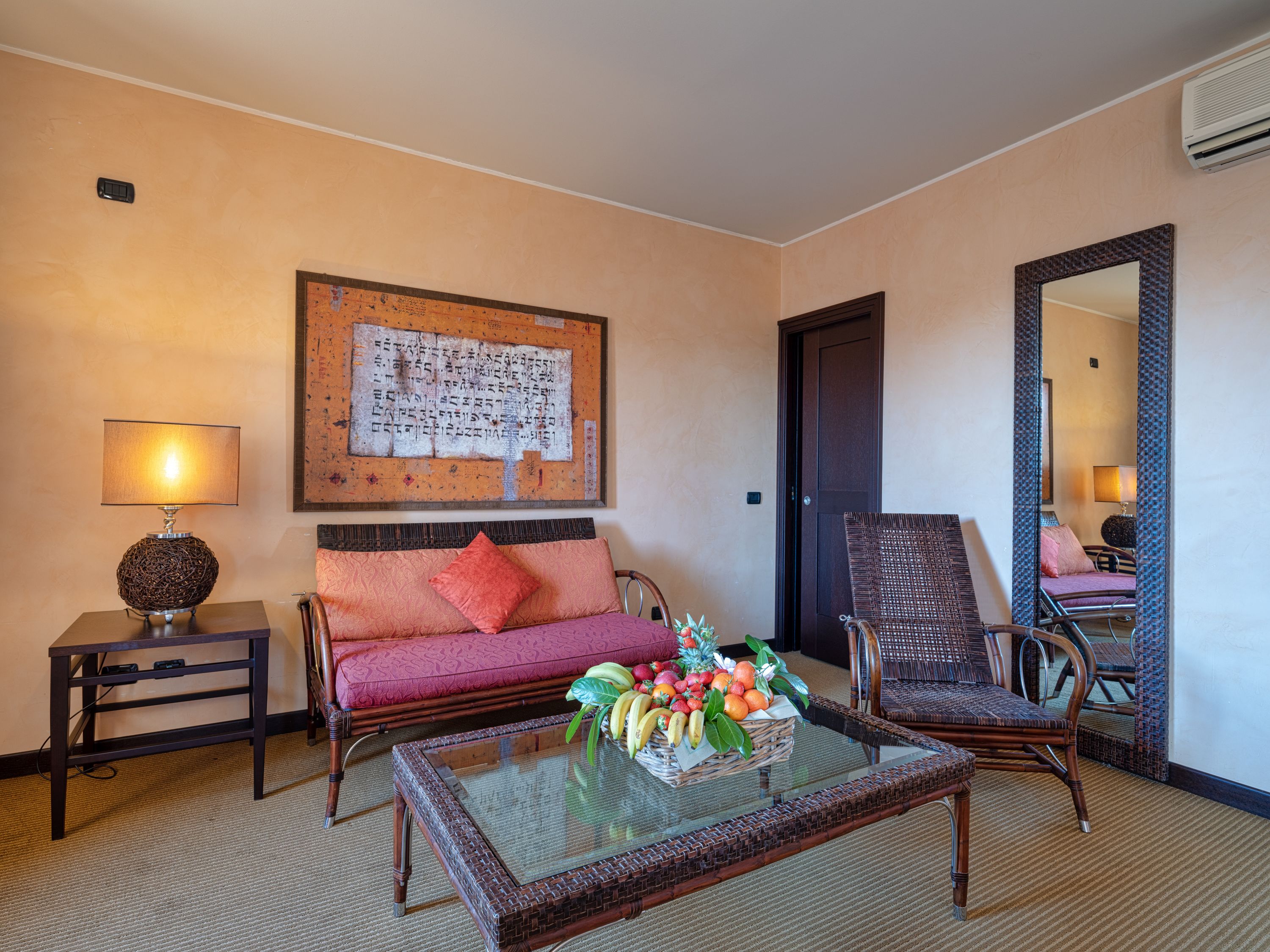 Une chambre d’hôtel avec un canapé, un fauteuil, une table basse en verre sur laquelle se trouvent des fruits, une lampe et une œuvre d’art accrochée au mur.