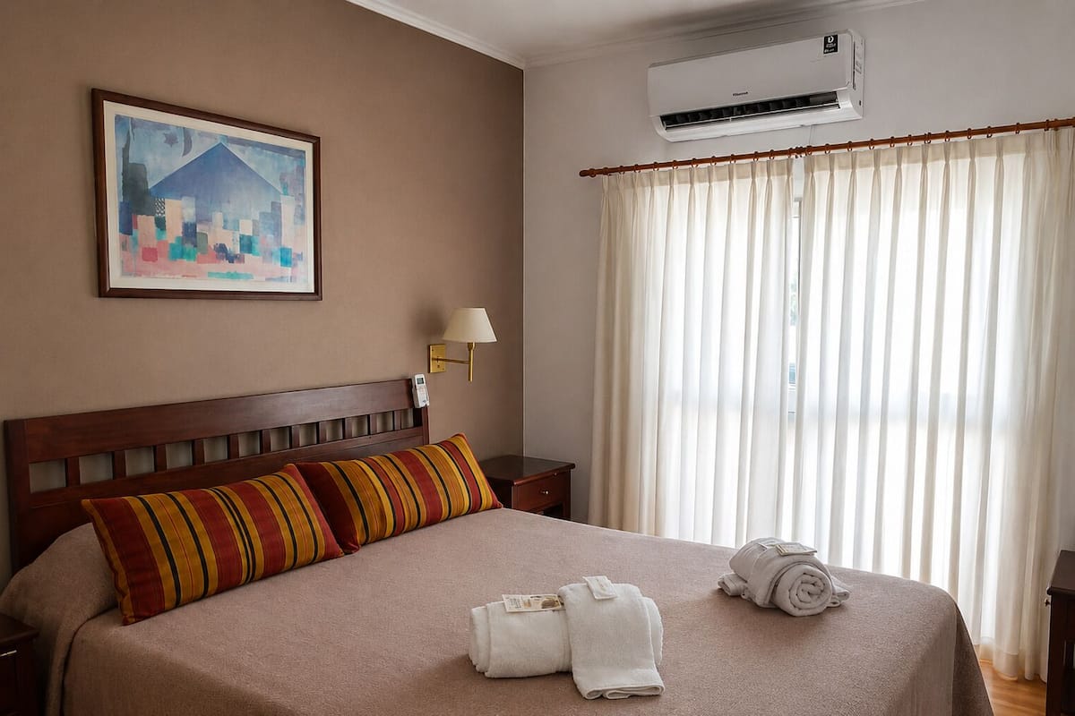 A hotel room with a bed, a painting on the wall, an air conditioner, and a window with curtains.