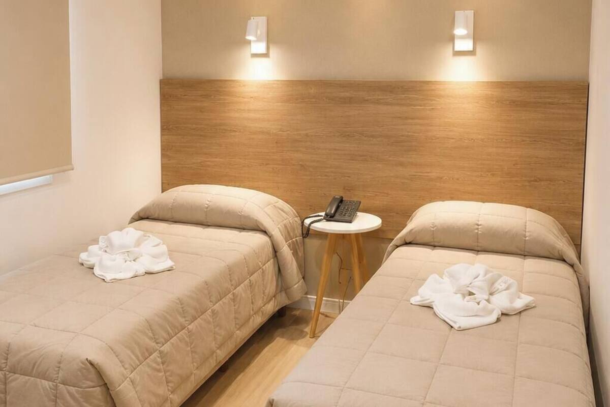 A hotel room with two beds, a small table, and two wall-mounted lights.