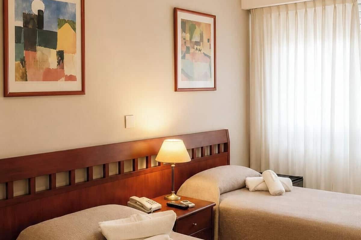 A hotel room with two beds, a wooden headboard, a bedside table with a lamp, a telephone, and two framed artworks on the wall.