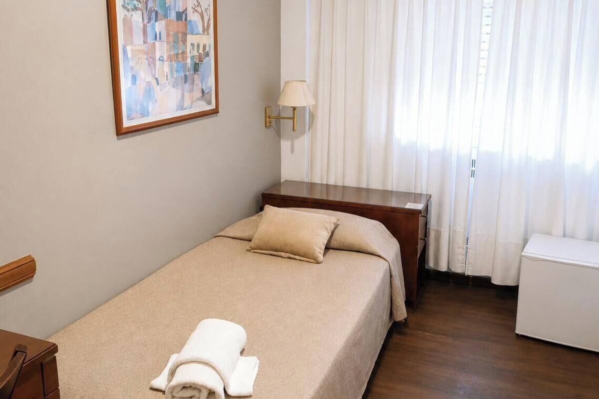 A hotel room with a bed, a desk, a chair, a lamp, a painting, and an air conditioning unit.