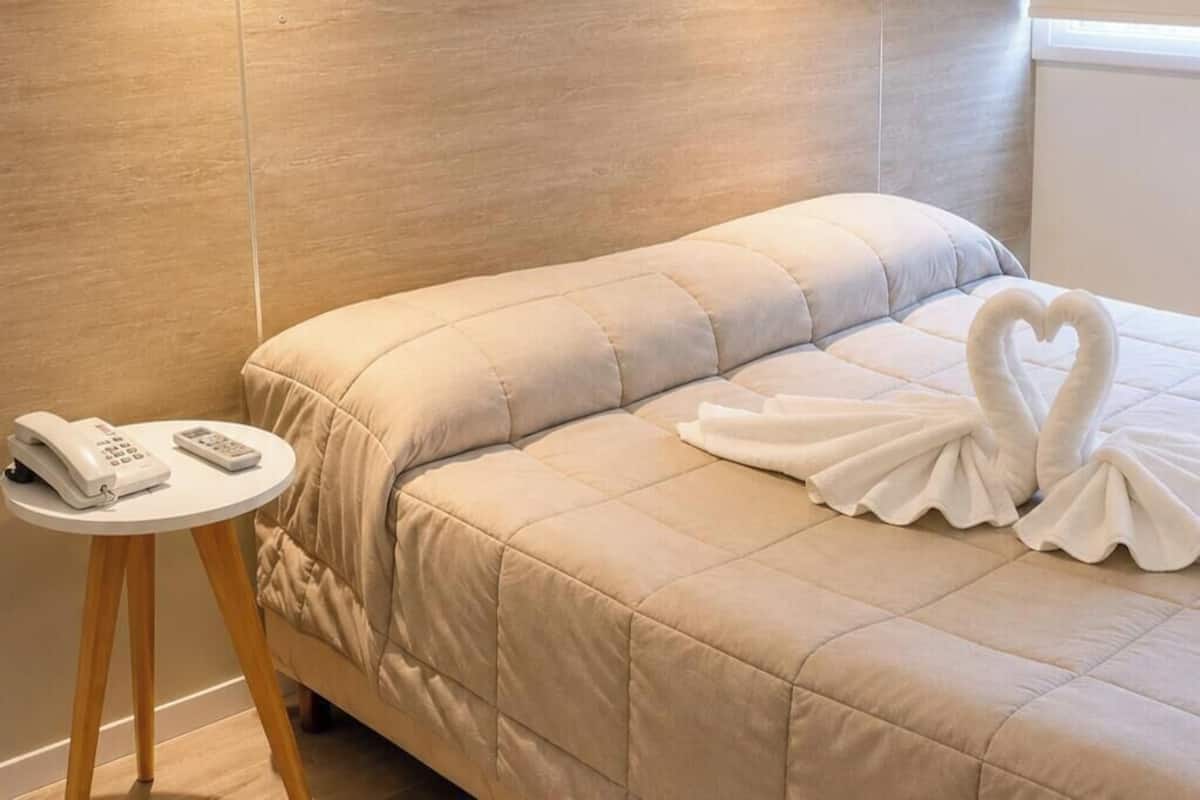 A hotel room with a bed, two wall-mounted lights, a bedside table with a phone and remote control, and a window with blinds.