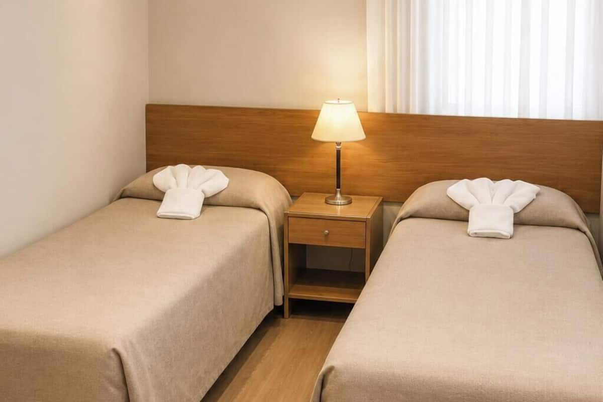 A hotel room with two single beds, a nightstand with a lamp, and a window with curtains.