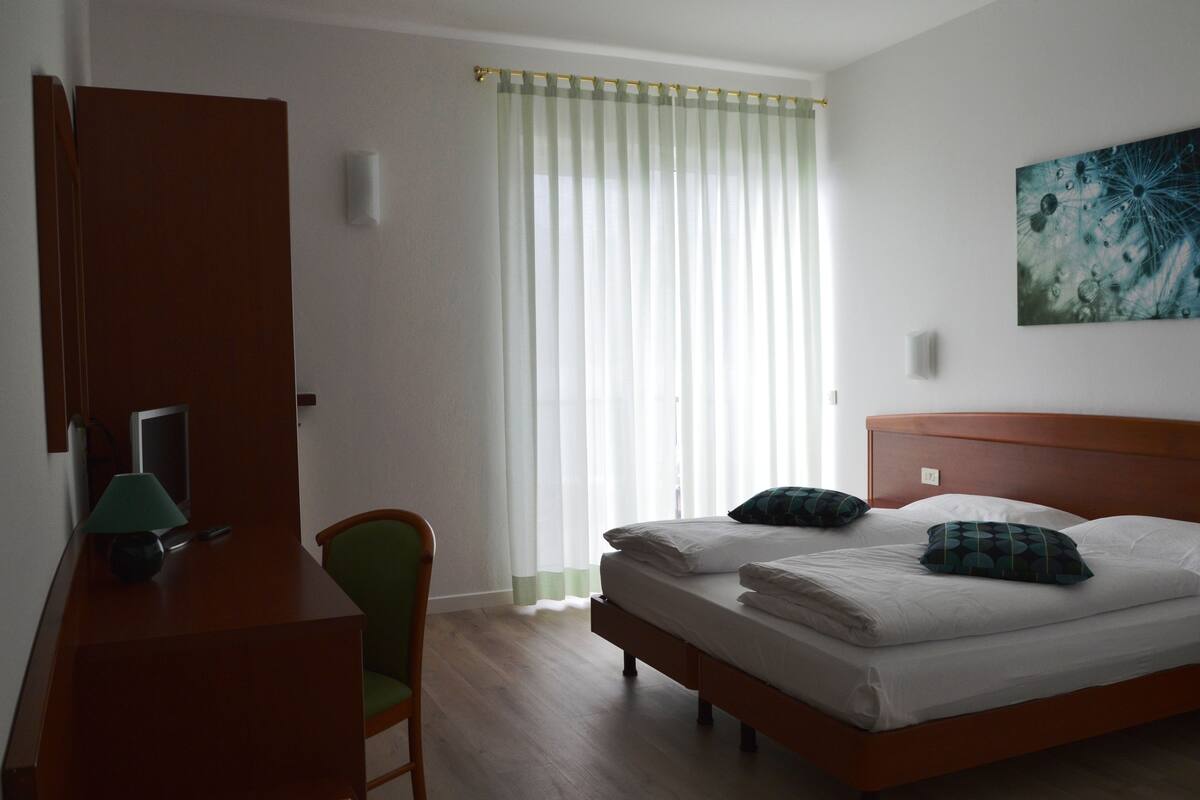 A bedroom with a wooden bed, a desk with a lamp, a chair, and a window with sheer curtains.