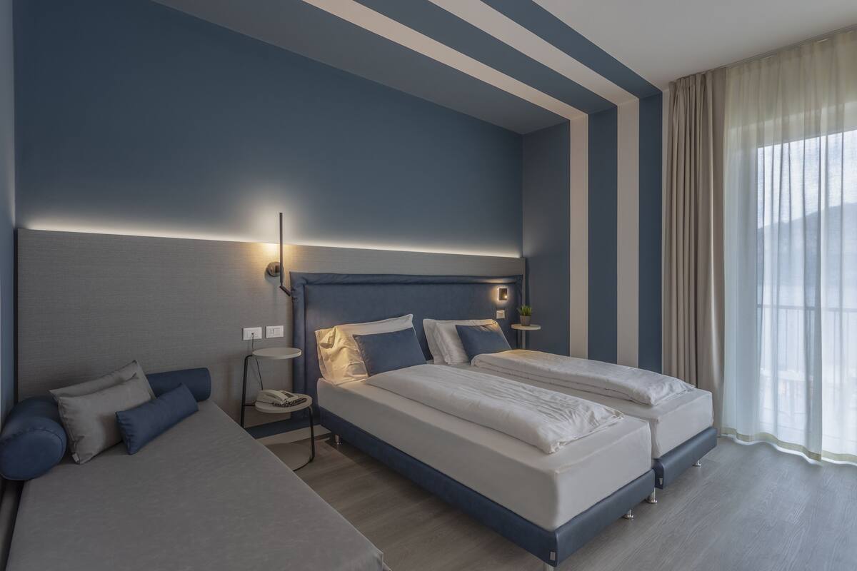 A modern hotel room with two beds, a bedside table with a lamp, and a large window with curtains.