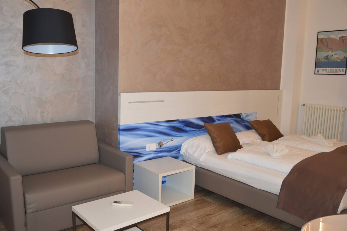 A modern hotel room with a sofa, a small table, a bed with a blue wall mural, and a lamp.