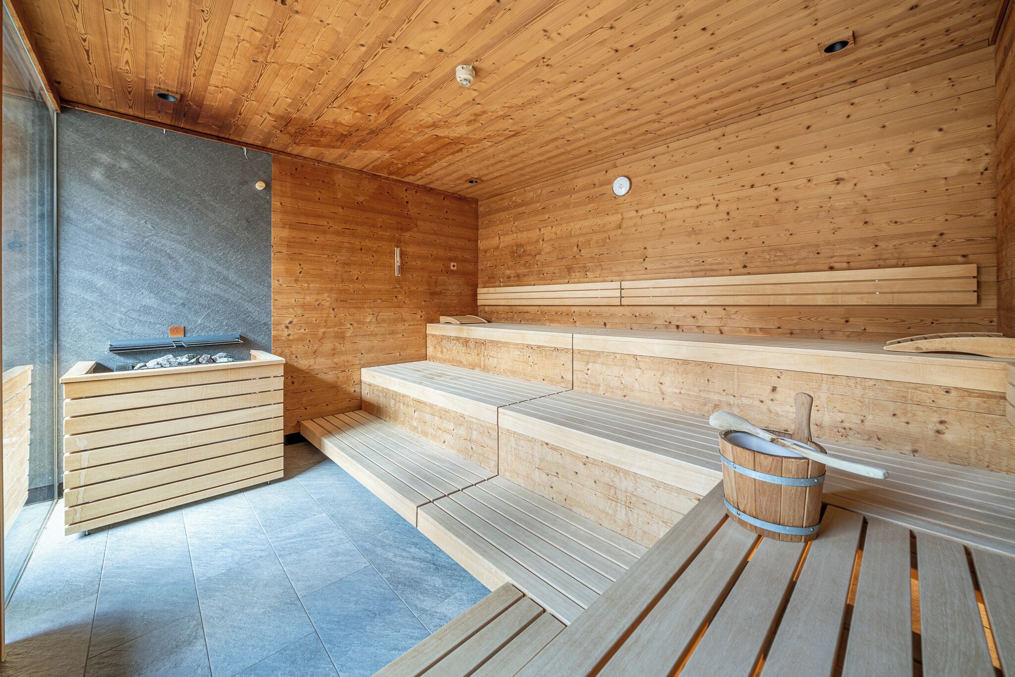 Un sauna avec des bancs en bois et un banc en pierre, un seau en bois avec une louche, et un mur en pierre.
