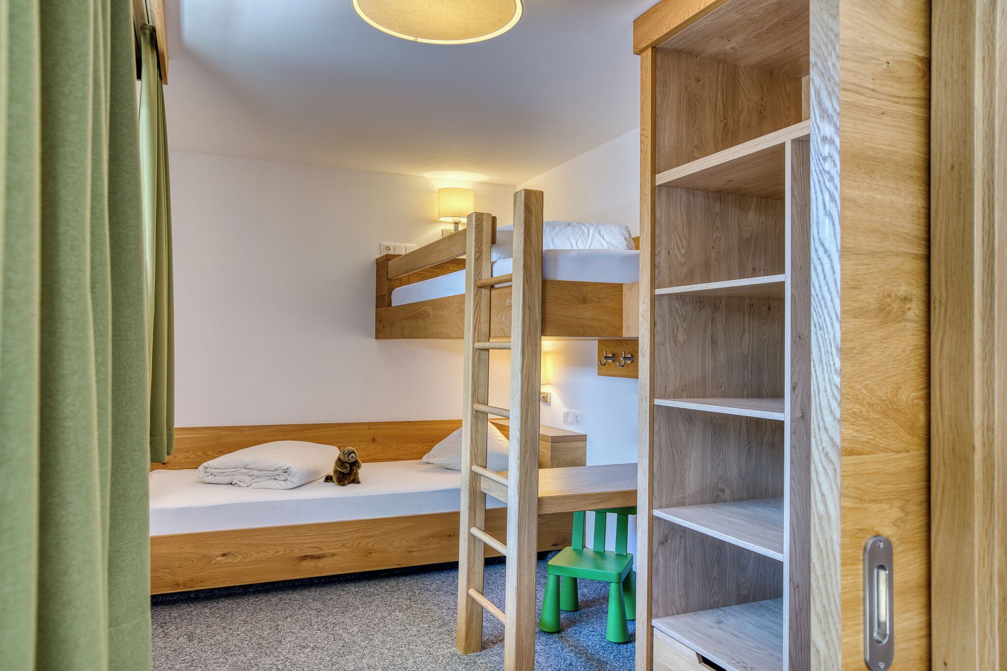 Cama litera con escalera, un escritorio, una silla y una estantería en una habitación con paredes blancas y una lámpara de techo.