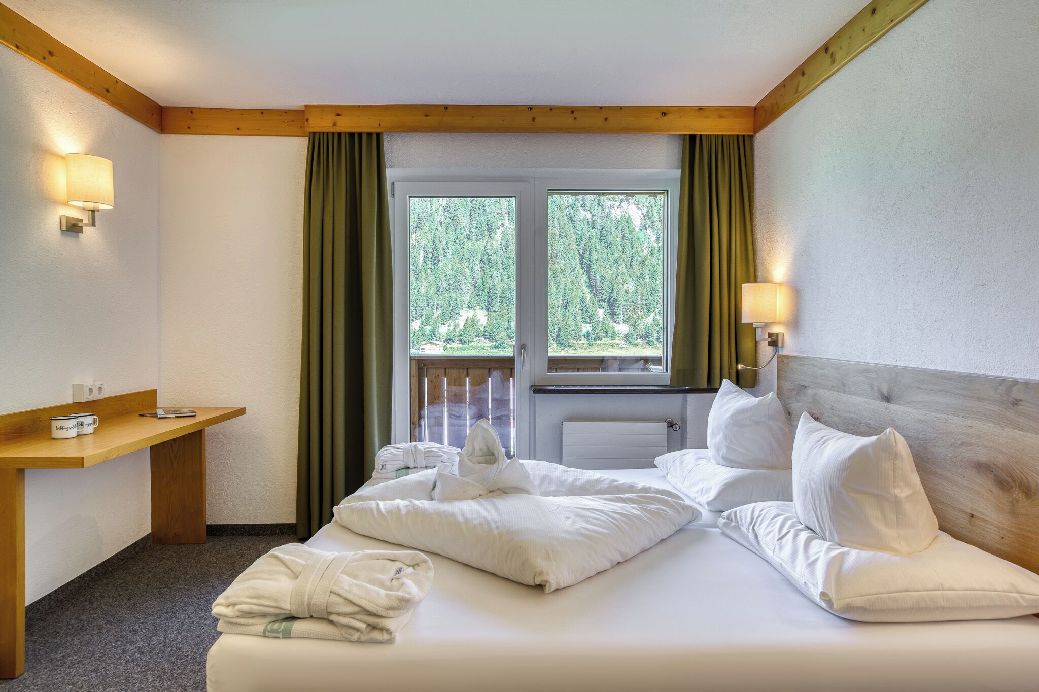 A hotel room with a bed, a wooden headboard, a nightstand with a lamp, a window with curtains, and a view of trees.