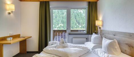 A hotel room with a bed, a wooden headboard, a nightstand with a lamp, a window with curtains, and a view of trees.