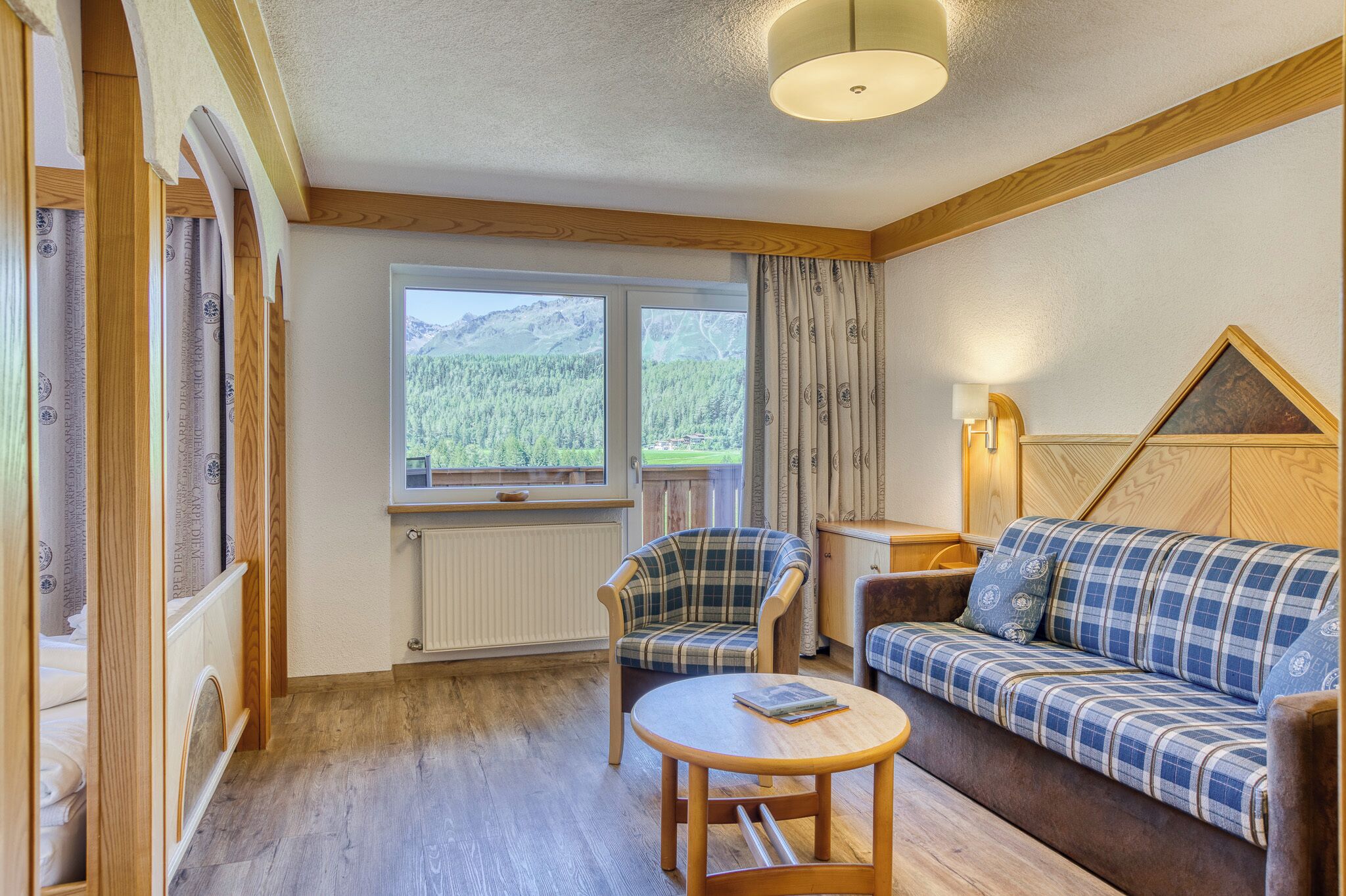A cozy room with a wooden floor, a plaid sofa, a chair, a small table, a window with a view of mountains, and a lamp.