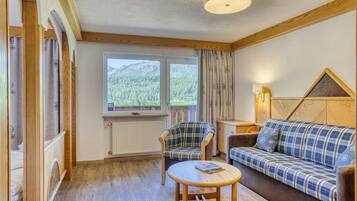 A cozy room with a wooden floor, a plaid sofa, a chair, a small table, a window with a view of mountains, and a lamp.