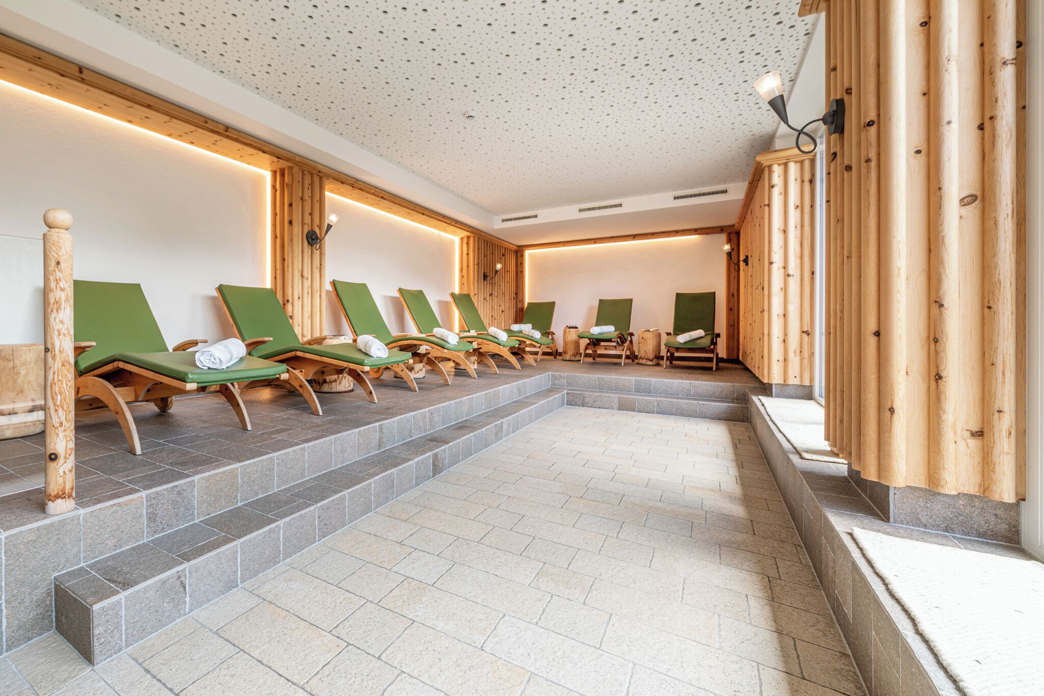Un área de spa con múltiples sillones reclinables verdes dispuestos en filas, paneles de madera en las paredes y un suelo de baldosas.