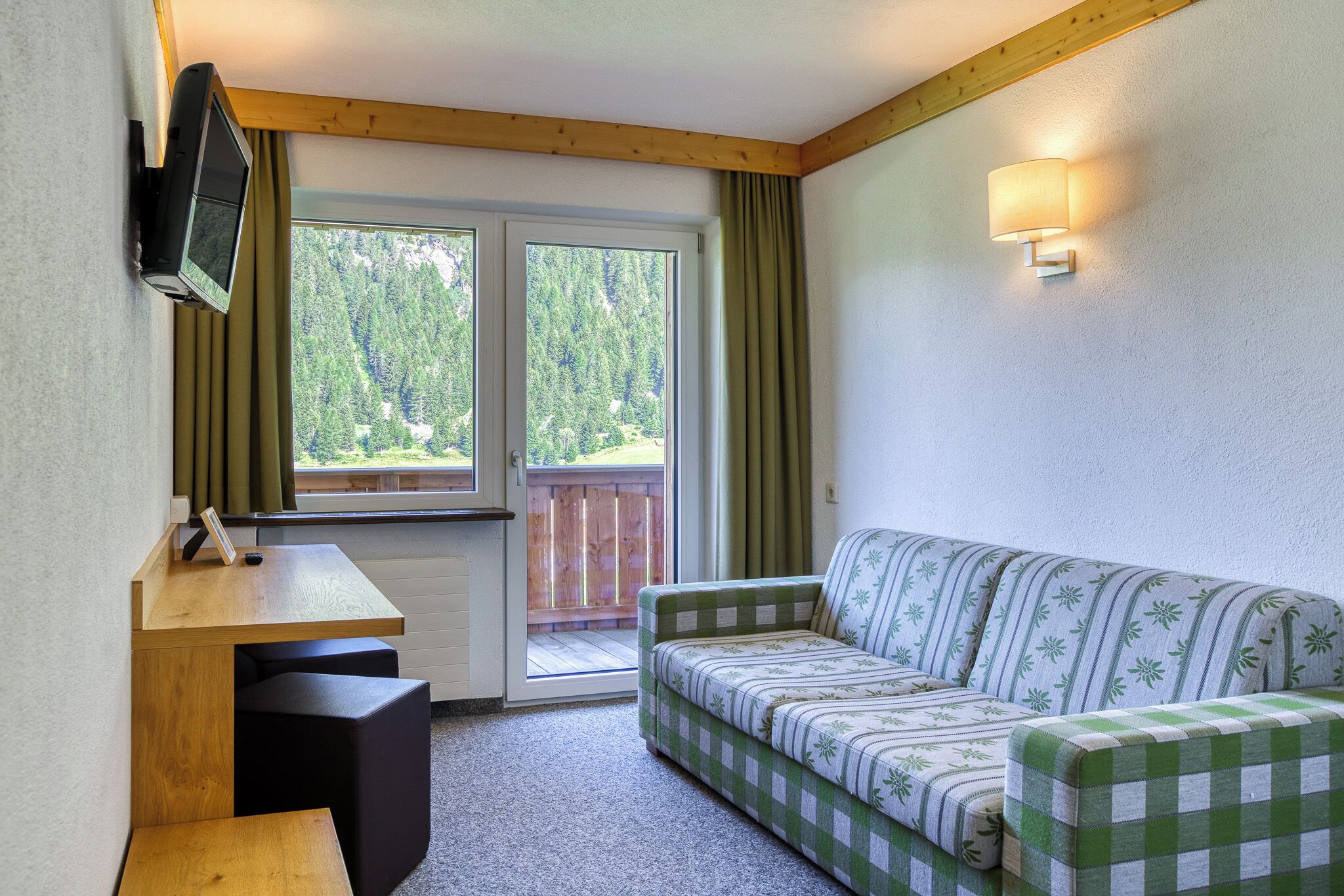 A room with a wooden desk, a green and white checkered sofa, a television mounted on the wall, and a sliding glass door leading to an outdoor area.