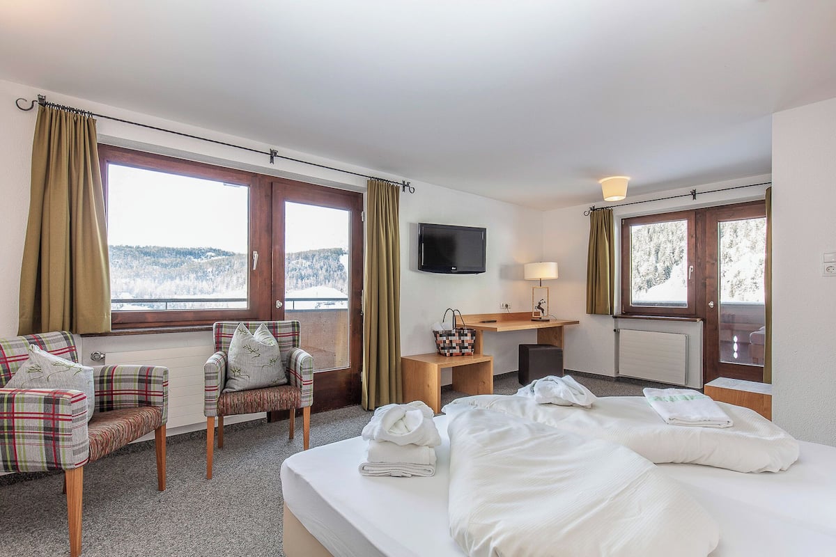 Habitación de hotel con cama, dos sillones, televisor, escritorio y ventanas amplias con cortinas.