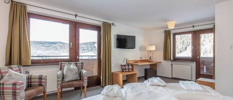 A hotel room with a bed, two chairs, a TV, a desk, and large windows with curtains.