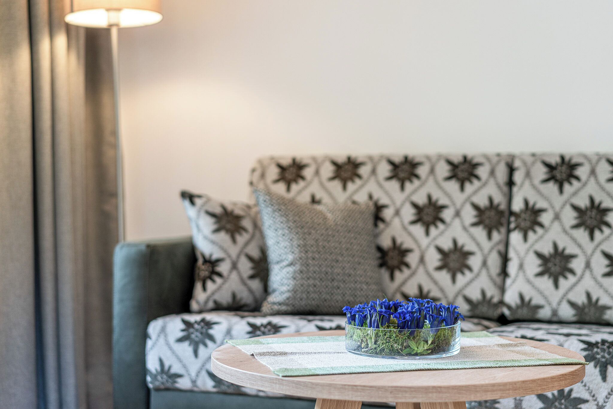 A sofa with a geometric pattern, a clear glass bowl with blue flowers on a wooden table, and a lamp in the background.
