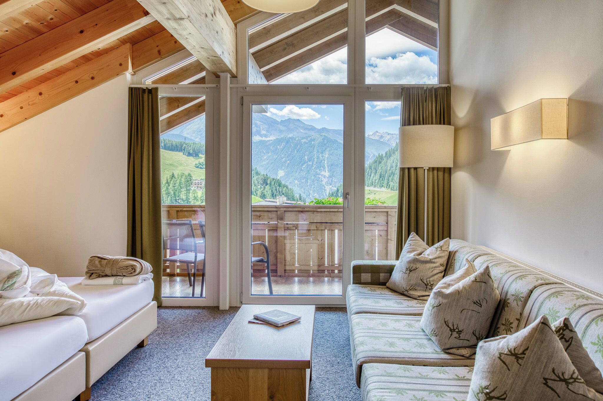 Ein Zimmer mit Bett, Sofa, Tisch und Blick auf Berge durch große Fenster.