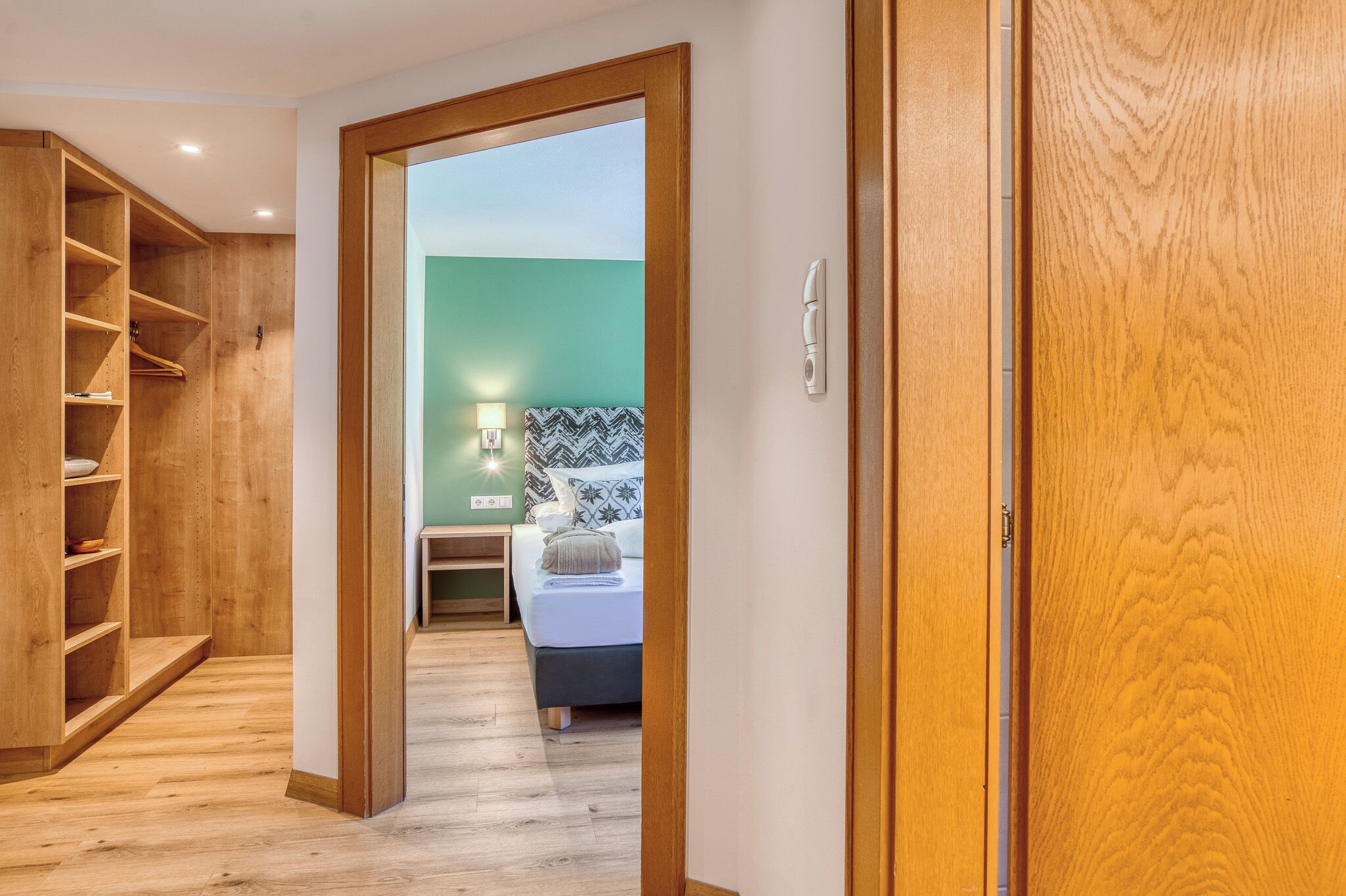 Un pasillo que conduce a un dormitorio con cama, mesita de noche y lámpara. A la derecha hay un armario de madera y, a la izquierda, una estantería de madera.