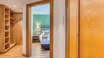 Un pasillo que conduce a un dormitorio con cama, mesita de noche y lámpara. A la derecha hay un armario de madera y, a la izquierda, una estantería de madera.