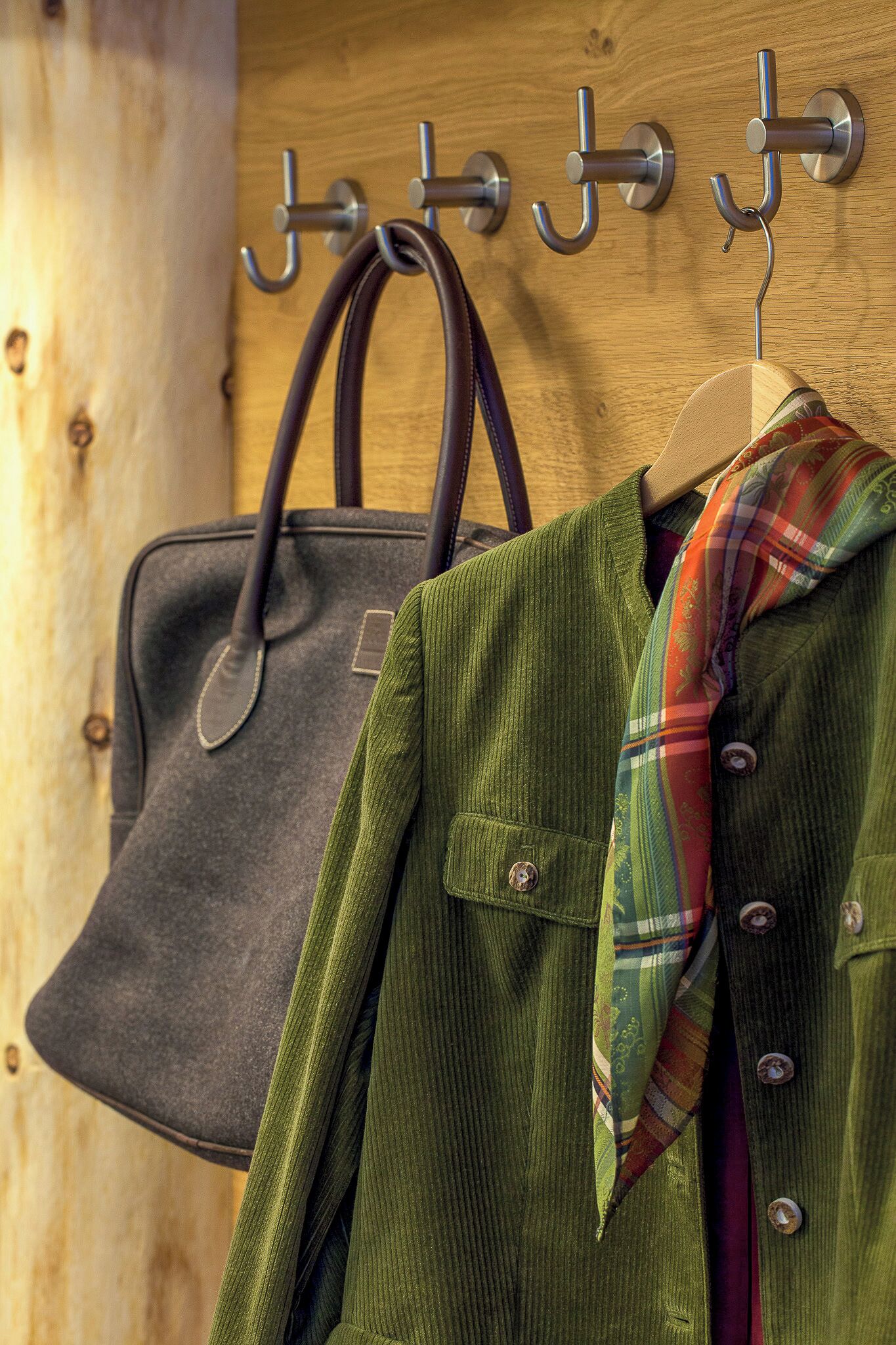 Una pared de madera con ganchos metálicos sosteniendo un bolso oscuro y una chaqueta verde con una bufanda de cuadros.