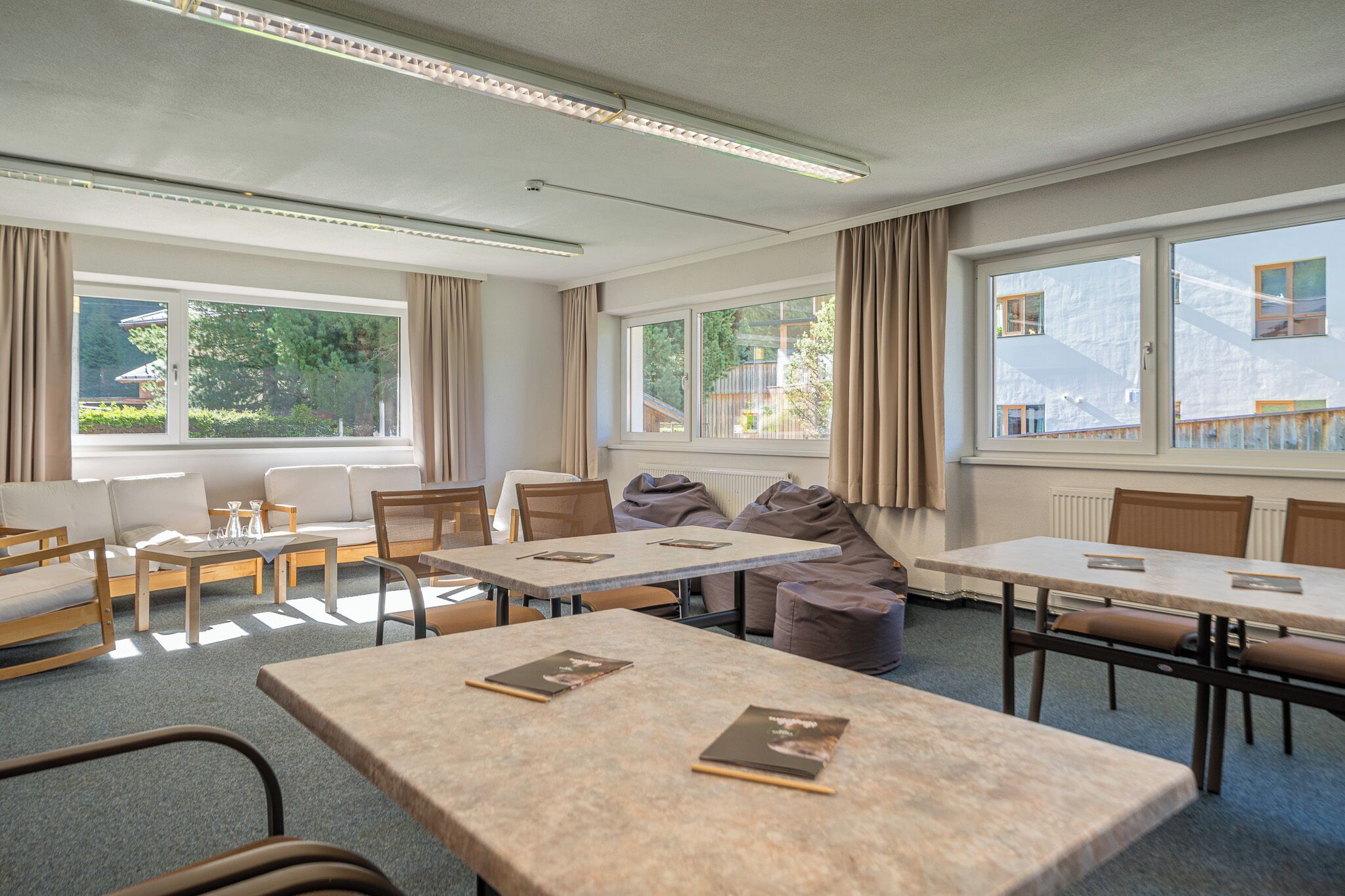 Habitación de hotel con sofá, dos mesas, sillas y ventanas grandes con cortinas.
