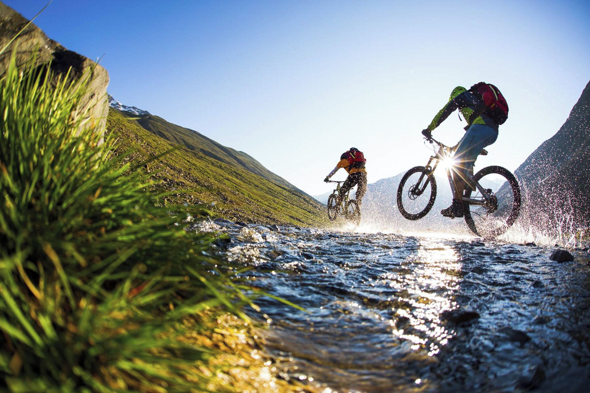 Dos ciclistas de montaña cruzando un arroyo en un entorno natural, con colinas al fondo.