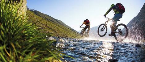 Dos ciclistas de montaña cruzando un arroyo en un entorno natural, con colinas al fondo.