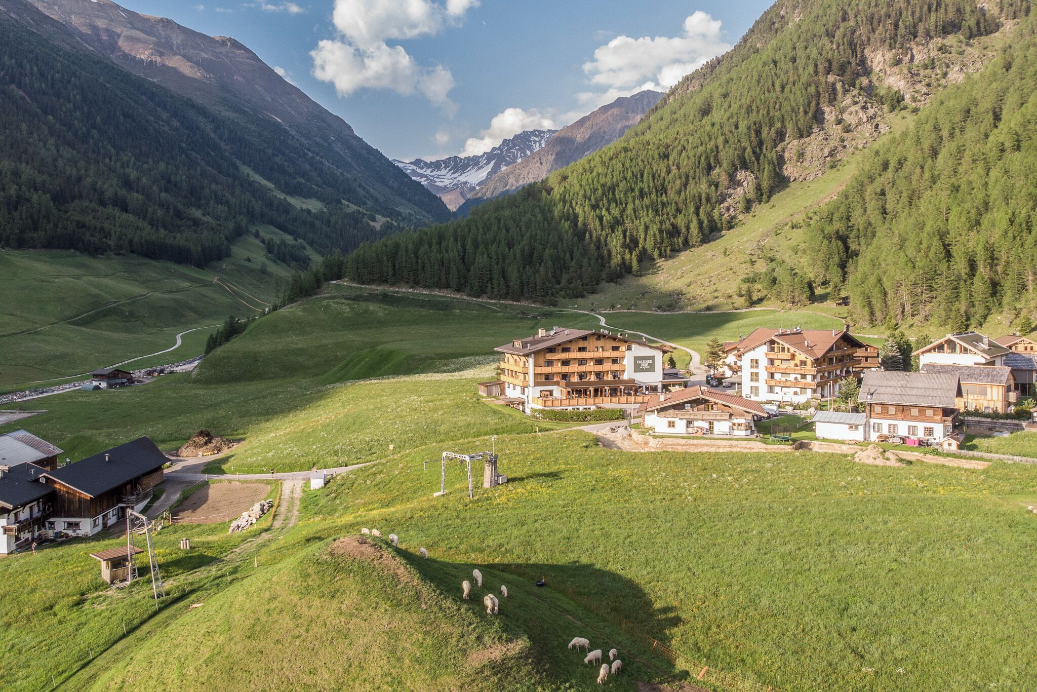 Un paisaje montañoso con un hotel que cuenta con varias construcciones, un telesilla y un rebaño de ovejas pastando.