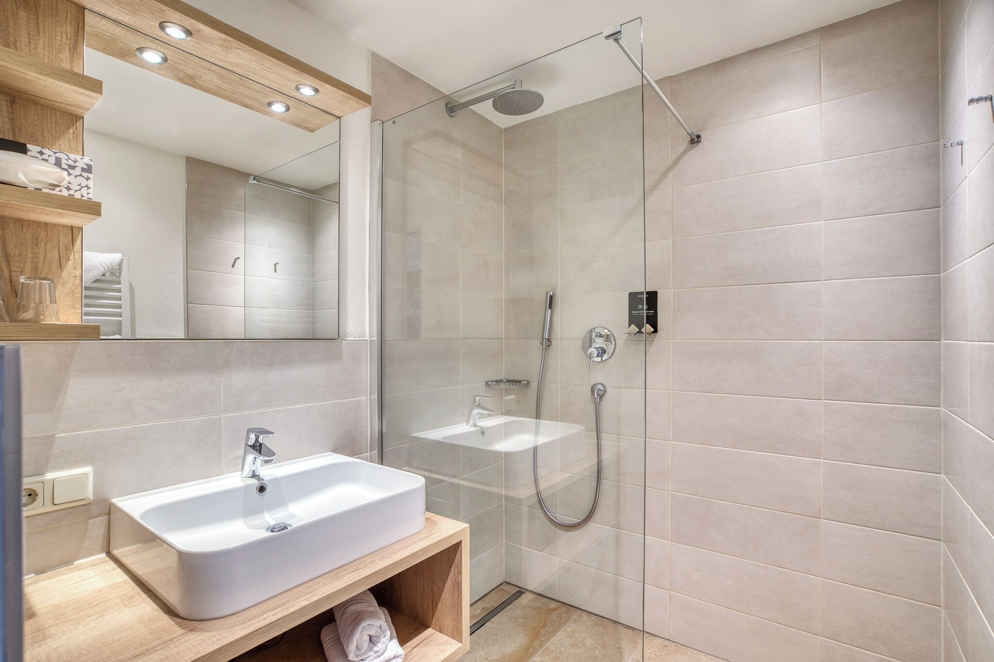 Un baño moderno con ducha envuelta en vidrio, un lavamanos blanco y un mueble de tocador de madera.
