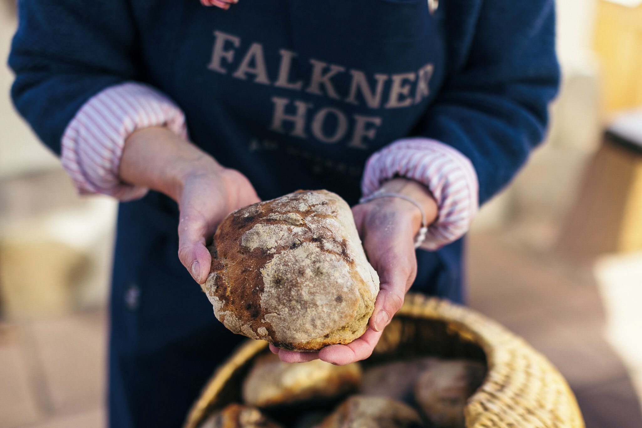 A person holding a loaf of bread, wearing a dark apron with 'FALKNER HOF' text.