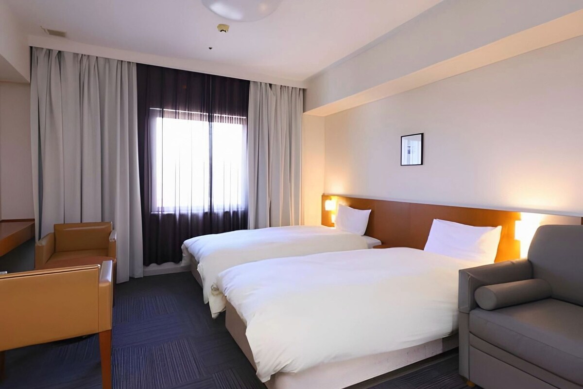Une chambre d’hôtel avec deux lits, un bureau, une chaise et une grande fenêtre avec des rideaux.