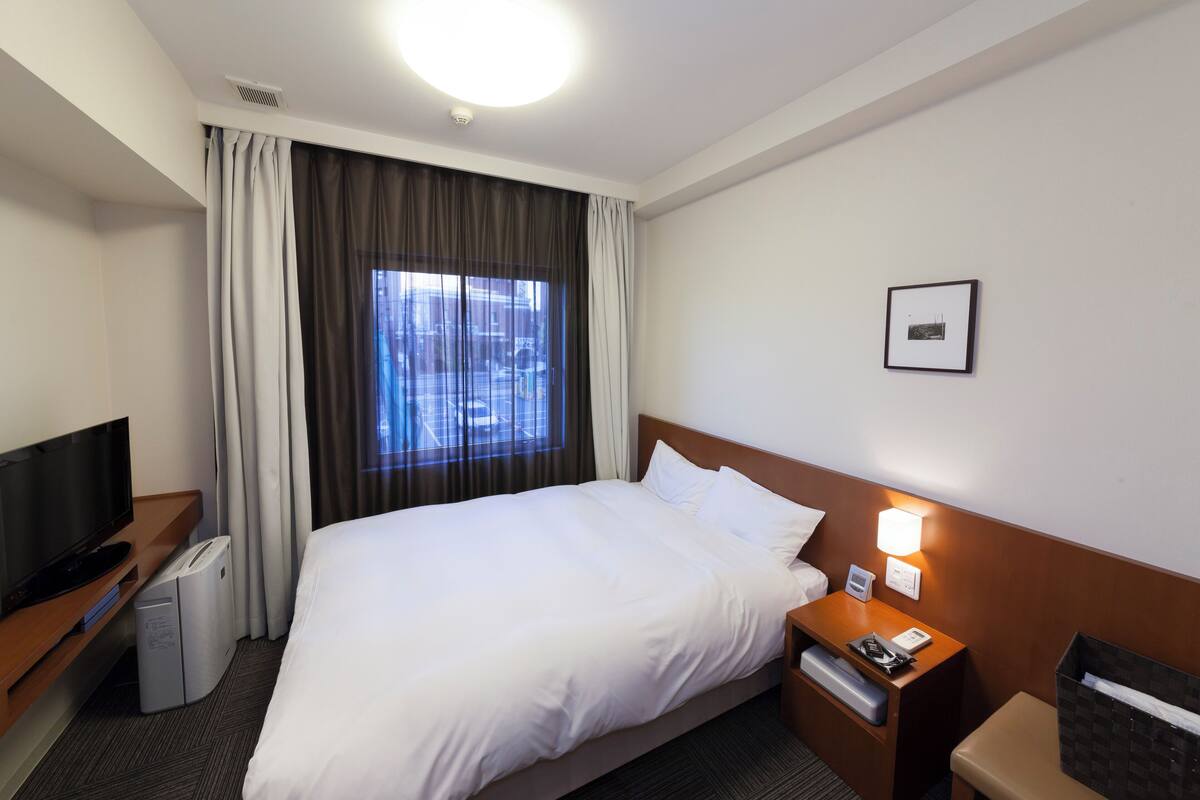 A hotel room with a bed, a TV, a desk, a nightstand, and a window with curtains.