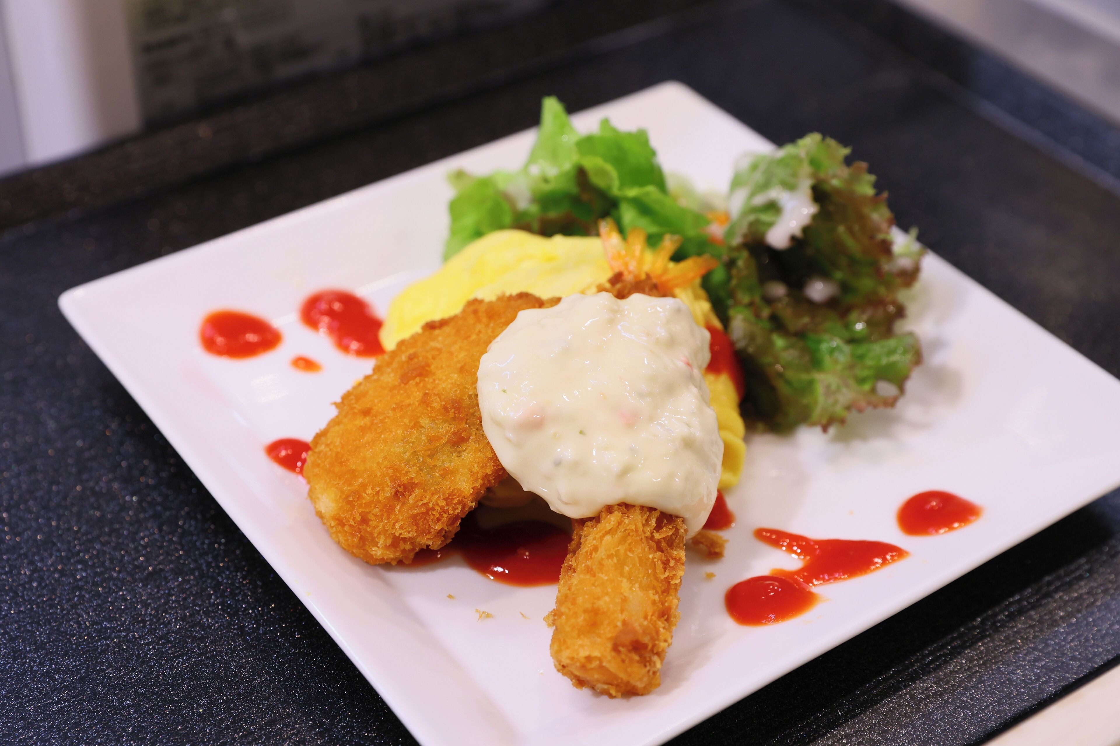 Un plato blanco rectangular con un alimento frito, una porción de salsa y una ensalada.