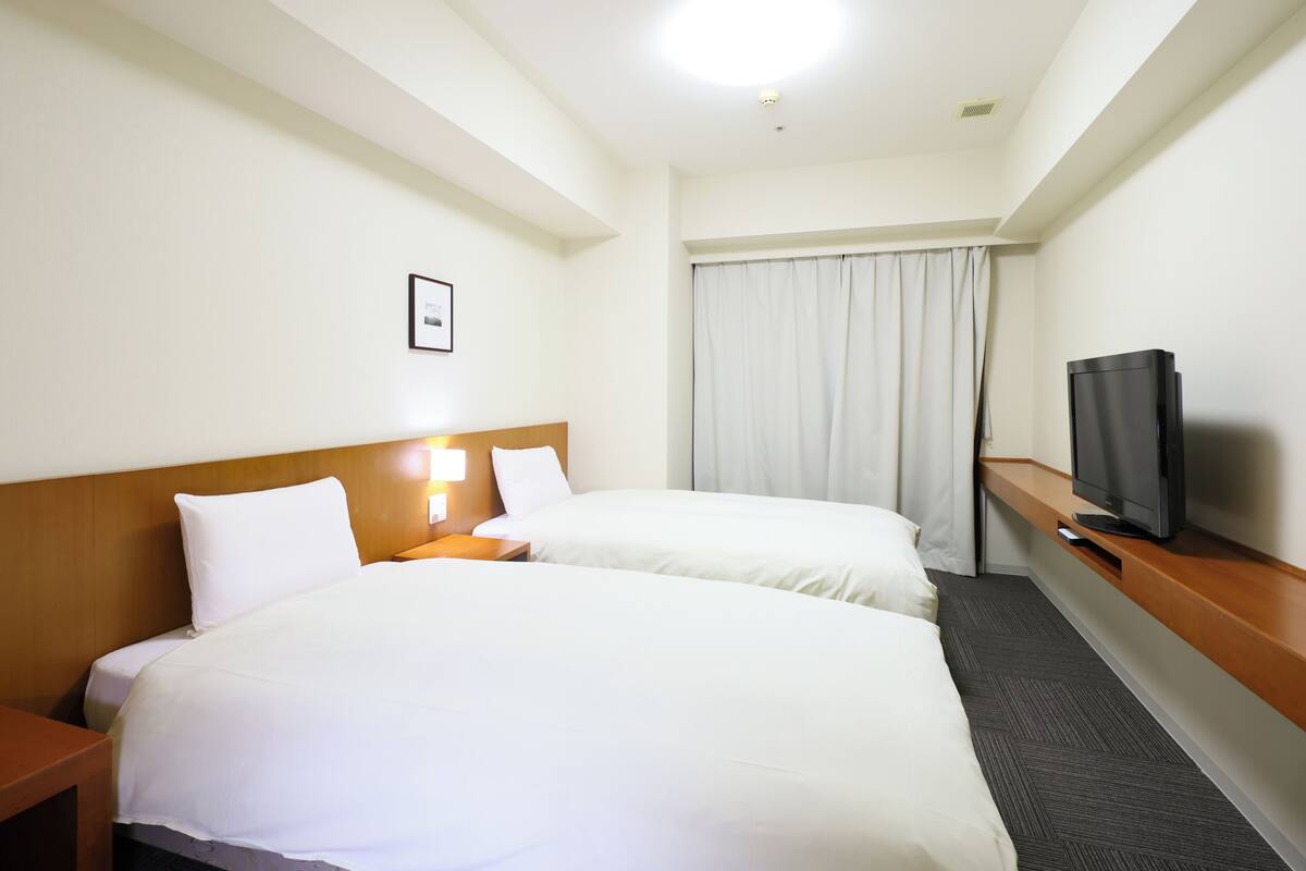 A hotel room with two beds, a TV, a nightstand, and a window with curtains.