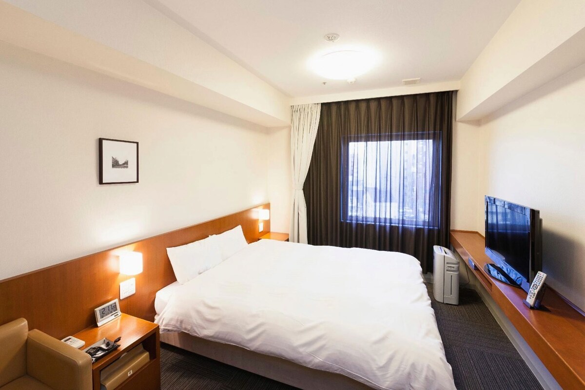 Une chambre d’hôtel avec un lit, une table de chevet, une télévision et une fenêtre avec des rideaux.